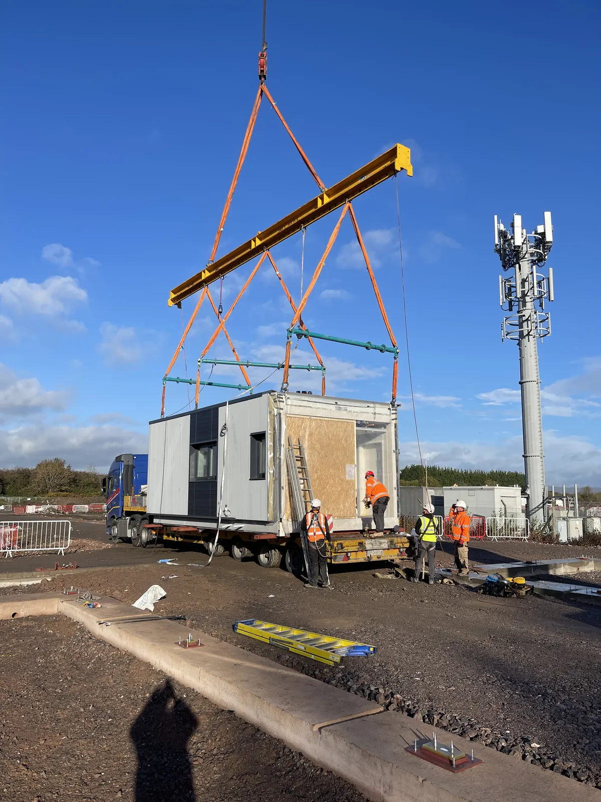 Craig Garbutt — MMC transport: volumetric unit being craned onto a trailer beside a telecoms mast ahead of site delivery